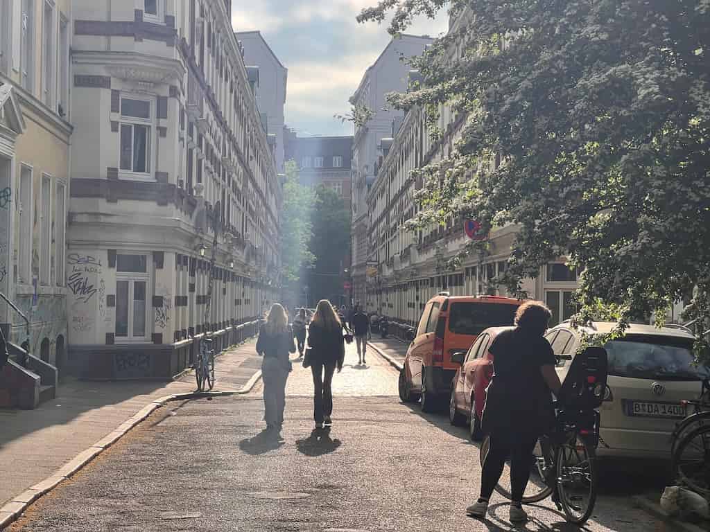 Eine ruhige Straße in einer Stadt, gesäumt von hohen, dekorativen Gebäuden mit weißen Fassaden und roten Backsteinakzenten. Links und rechts stehen geparkte Autos. Einige Menschen gehen oder fahren mit dem Fahrrad auf der Straße. Die Sonne scheint, und es gibt einige Schatten auf der Straße.